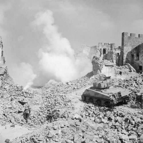 A Sherman tank advances through the rubble and smoke of the Monte Cassino battlefield, supported by New Zealand infantry.