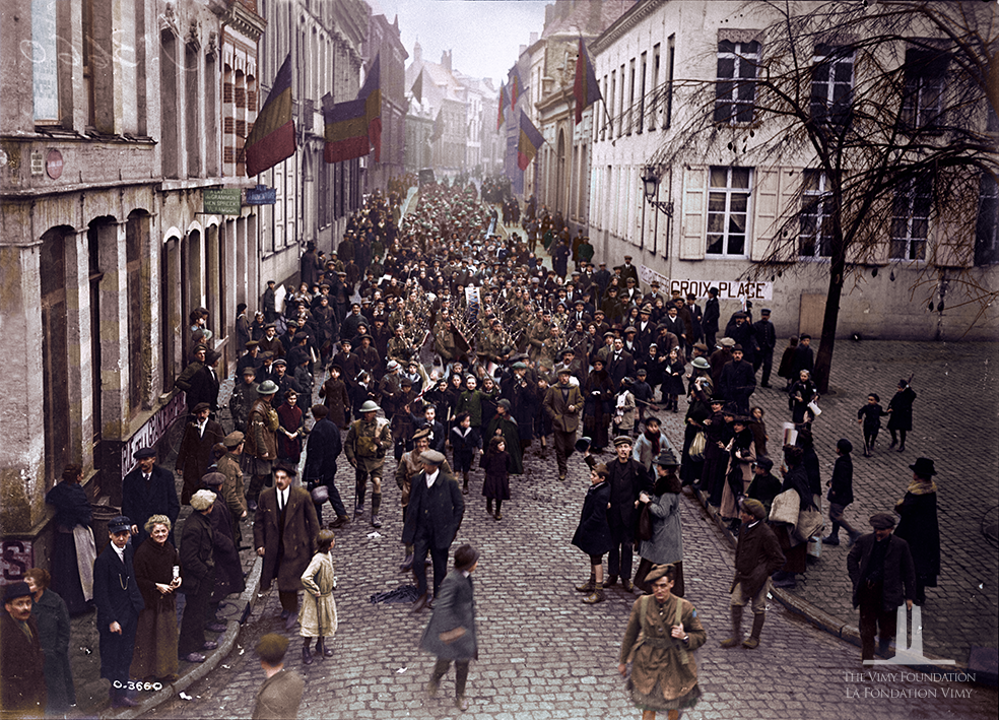 Colourised photo of Canadian troops marching through Mons. They are surrounded by grateful French citizens. French tricolour flags can be seen flying from the white buildings that flank the boulevard up which the Canadians are walking.