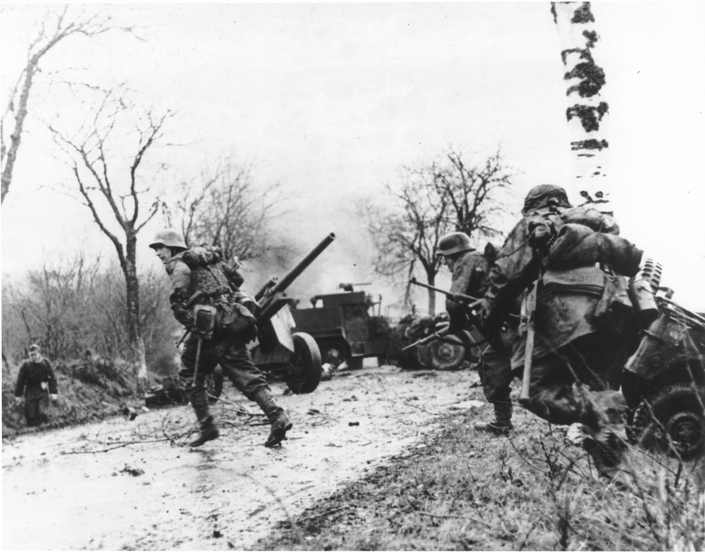 Wehrmacht soldiers crossing a road passed knocked out American artillery pieces and vehicles at the Battle of the Bulge